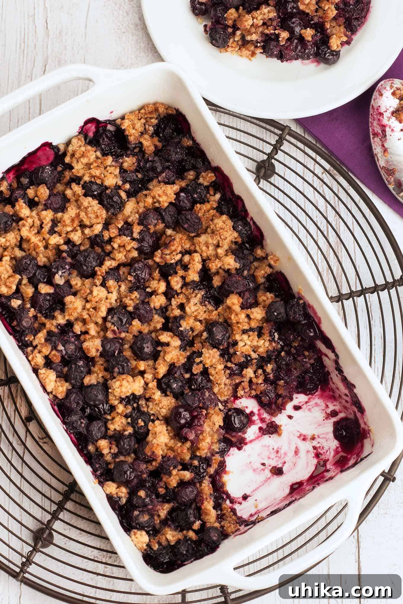 Blueberry Bliss with a Cornflake Crunch 2 Freshly baked blueberry crisp in a baking dish with a serving removed and placed on a plate to the side. Golden, crispy topping and bubbling, juicy blueberries are visible.