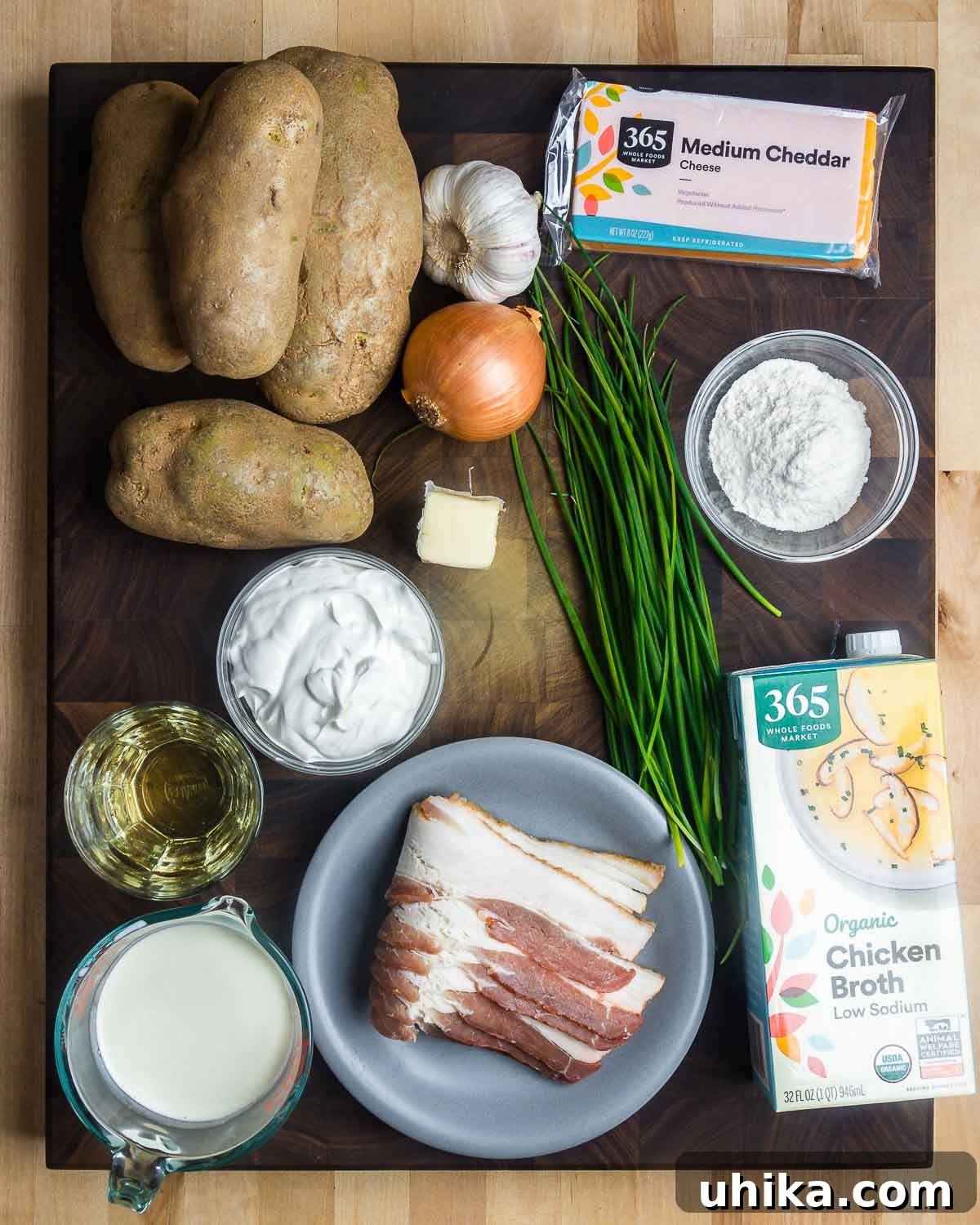 Hearty Loaded Potato Soup 3 Ingredients shown: russet potatoes, garlic, onion, shredded cheddar cheese, fresh scallions/chives, all-purpose flour, dry white wine, sour cream, thick-cut bacon, heavy cream, and low-sodium chicken broth.