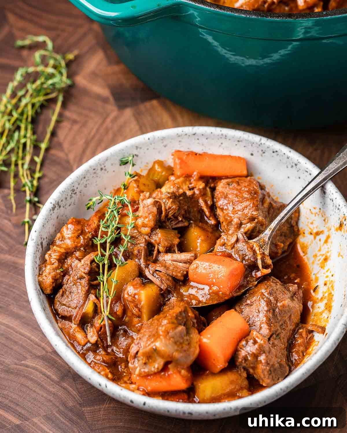 Rich Guinness Beef Stew 11 A beautiful white bowl filled with Guinness beef stew, garnished with fresh herbs, with a rustic blue Dutch oven pot steaming in the soft-focus background, highlighting the warmth and richness of the dish.
