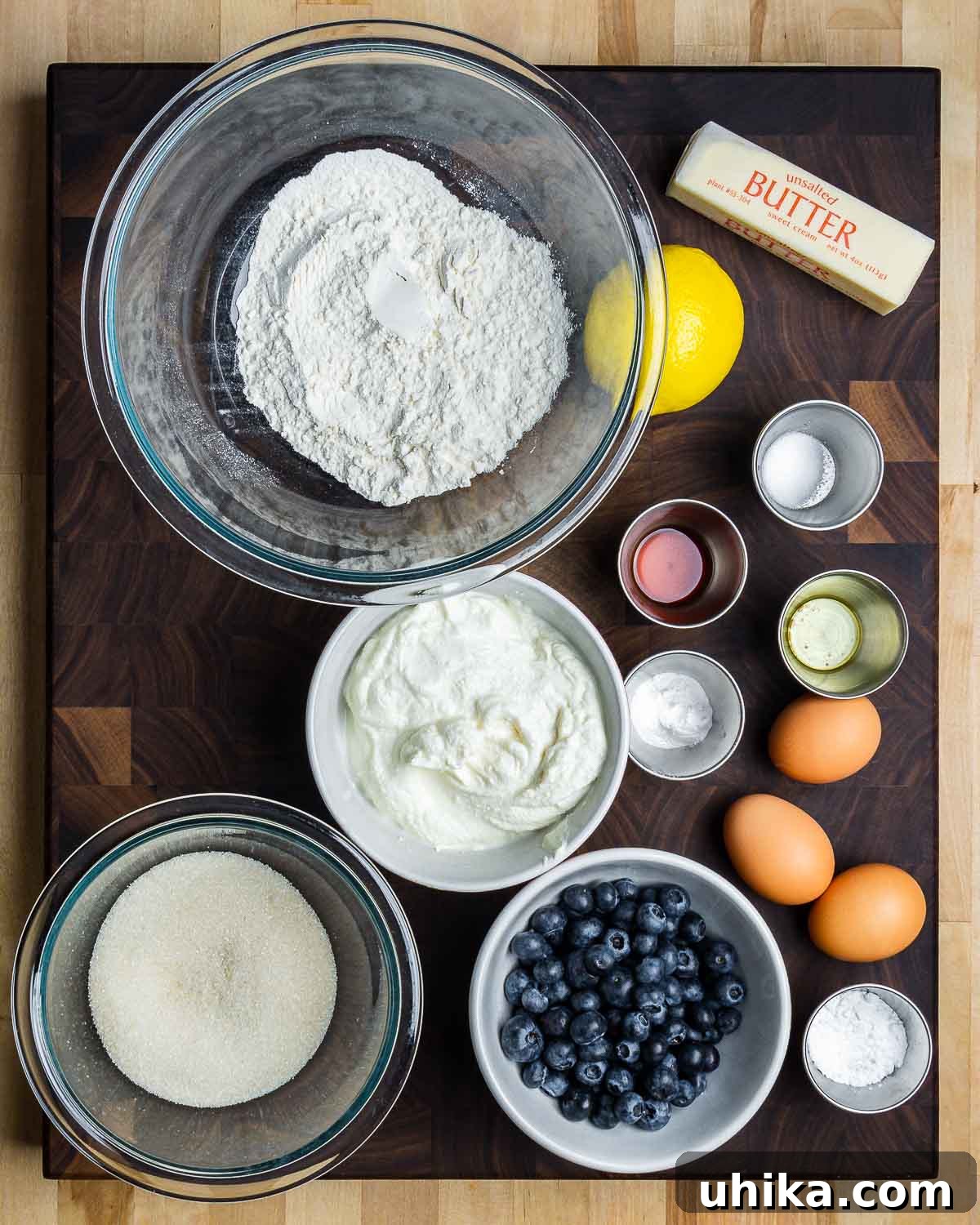 Overhead shot of ingredients laid out for lemon blueberry ricotta cake recipe, including lemons, blueberries, eggs, flour, and a whisk.