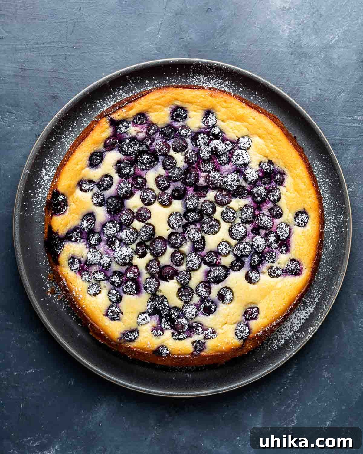 Overhead shot of a full lemon blueberry ricotta cake on a grey plate, placed on a blue board.
