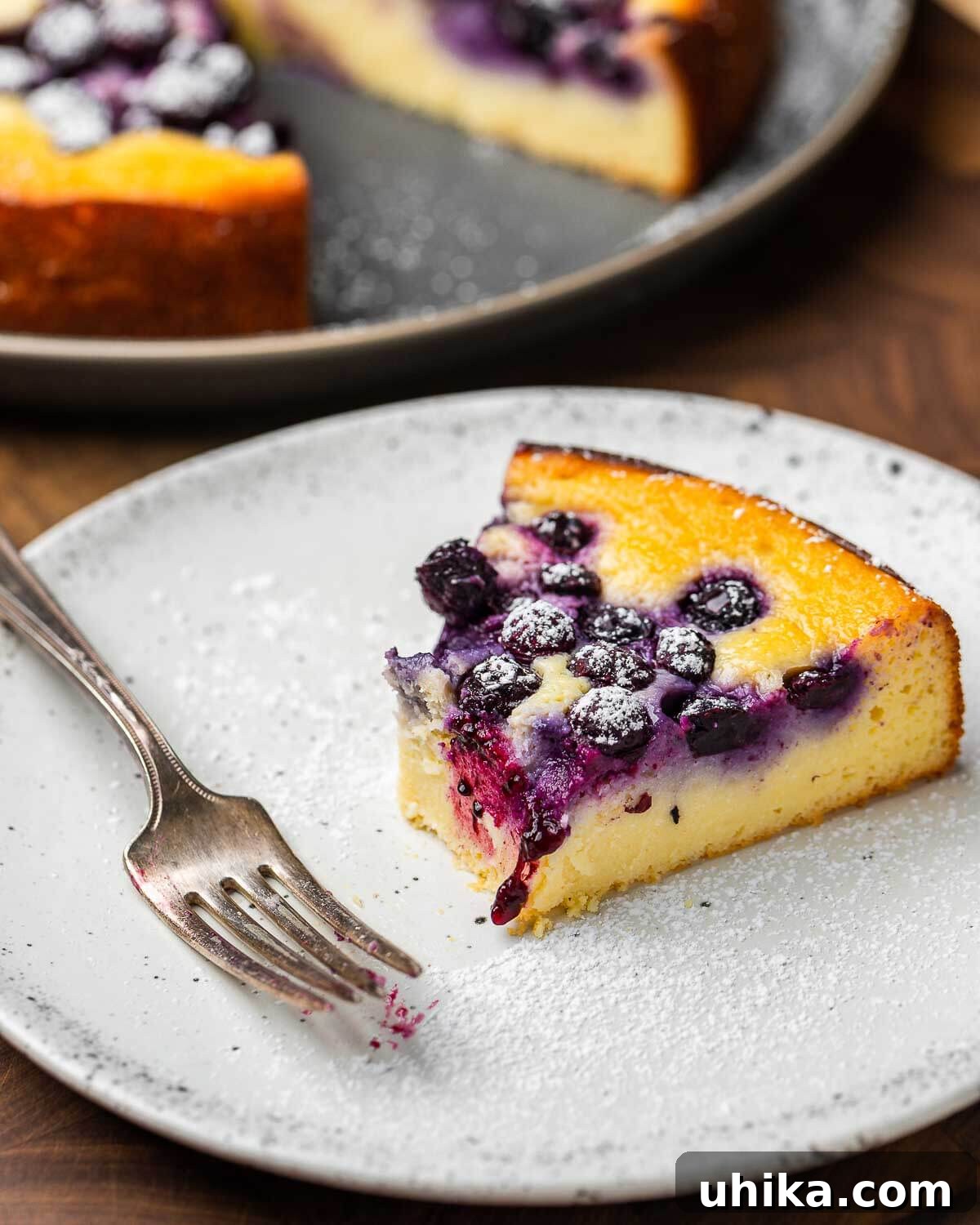 A bite taken out of a slice of lemon blueberry ricotta cake on a white plate, highlighting its moist texture and blueberries.