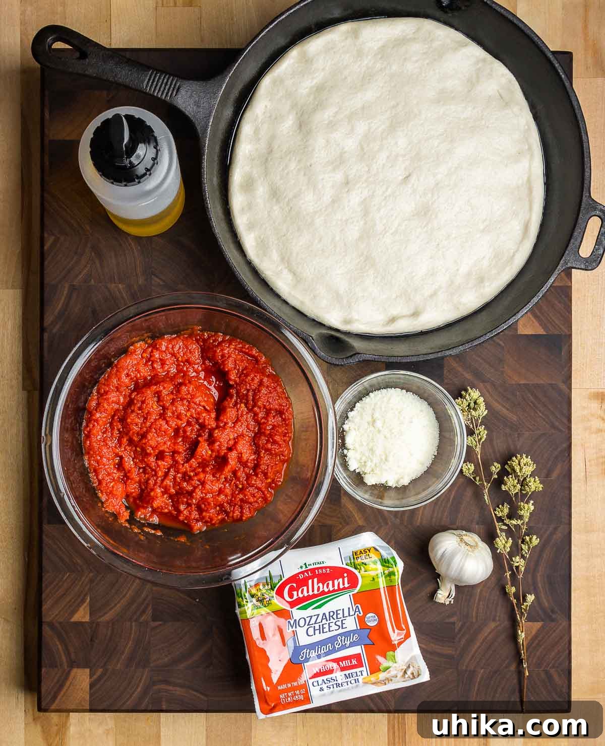 Skillet Pizza Perfection 3 All ingredients laid out for cast iron pan pizza, including pizza dough in a pan, olive oil, crushed tomatoes, Pecorino, mozzarella, garlic, and Sicilian oregano.