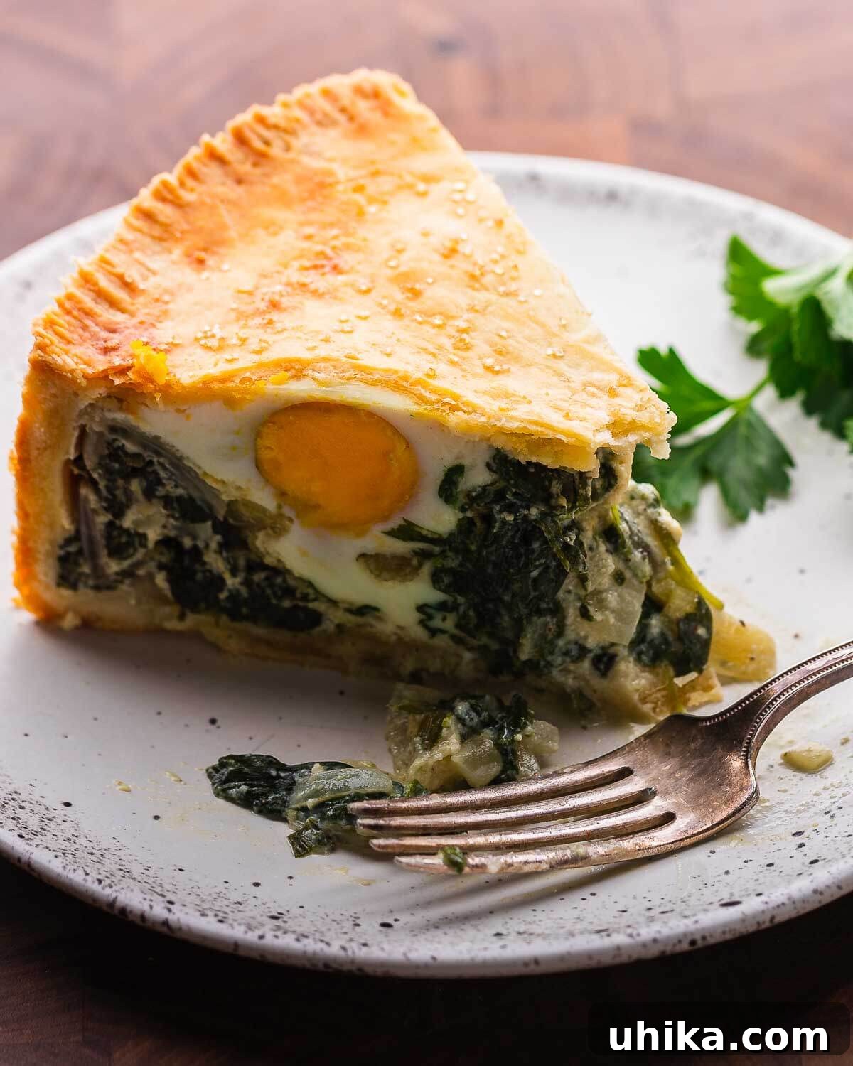 Classic Italian Easter Pie 2 A perfectly sliced piece of Torta Pasqualina, showcasing layers of golden crust, vibrant green filling, and a visible whole egg, presented on a white plate with a fork.