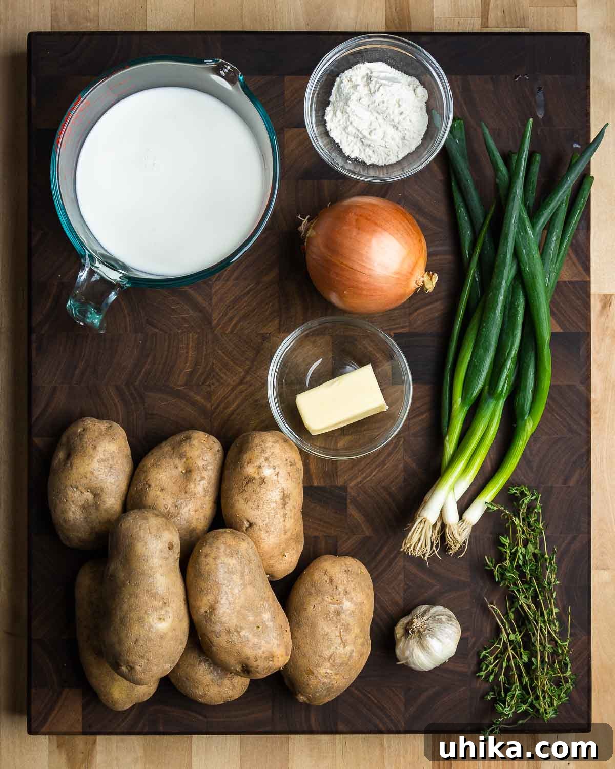Ultimate Creamy Potato Gratin 3 A flat lay photograph showcasing the fresh ingredients required for scalloped potatoes: milk, flour, green onions, white onion, butter, russet potatoes, garlic cloves, and fresh thyme sprigs.