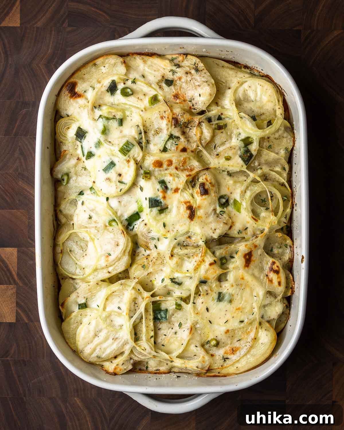 Ultimate Creamy Potato Gratin 7 An overhead shot of a baking dish filled with golden-brown scalloped potatoes on a dark walnut cutting board, ready for serving.