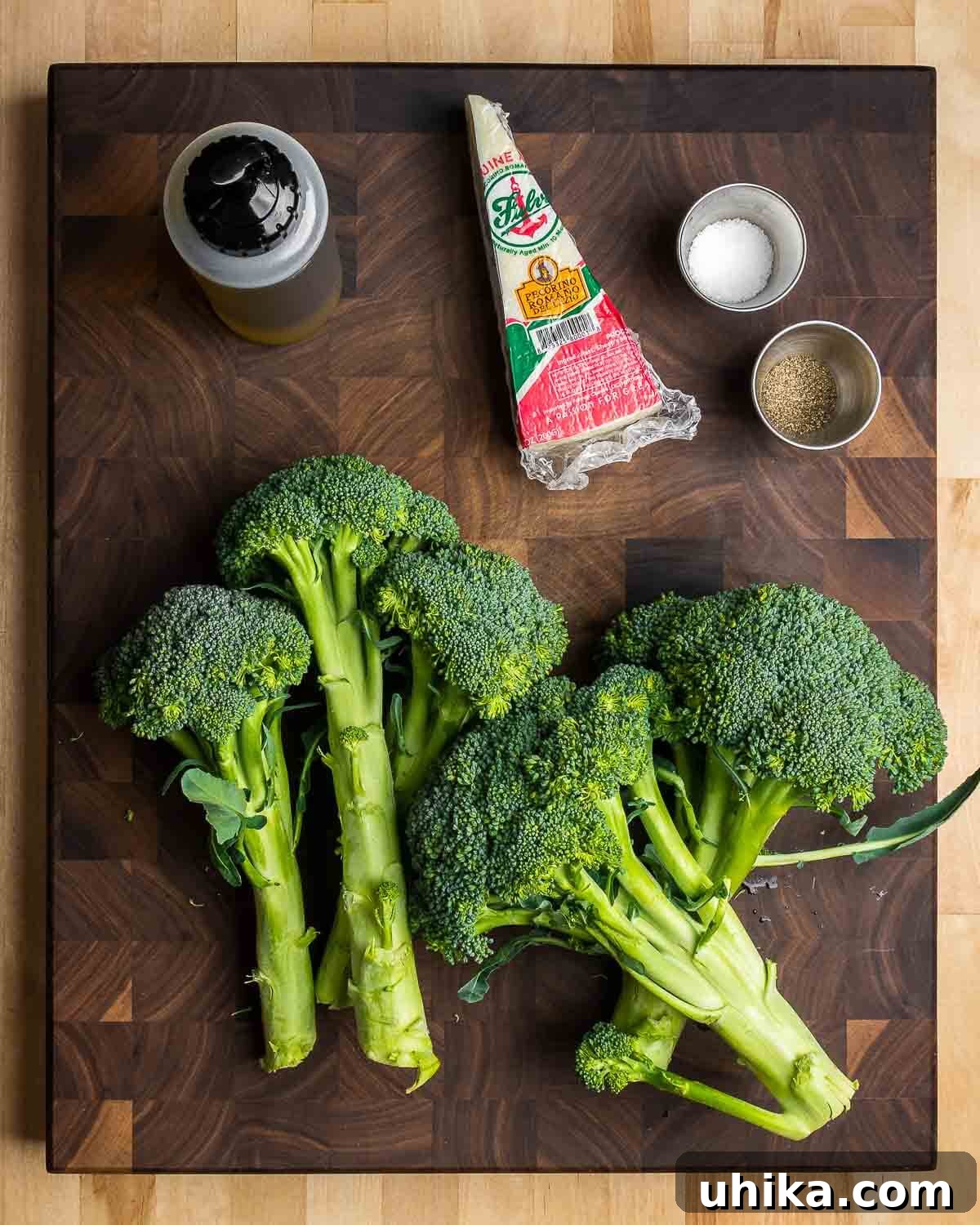 Crispy Roasted Broccoli 3 A flat lay photograph showcasing the essential ingredients for roasted broccoli: fresh broccoli florets, a bottle of extra virgin olive oil, a wedge of Pecorino Romano cheese, a bowl of kosher salt, and black peppercorns.