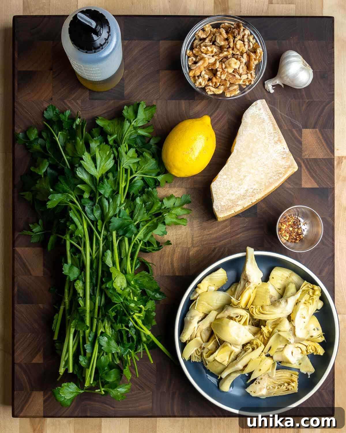 Vibrant Artichoke Pesto 3 Ingredients for artichoke pesto: extra virgin olive oil, walnuts, garlic cloves, fresh parsley, bright lemons, Parmigiano Reggiano cheese block, and dried hot red pepper flakes, all arranged around a bowl of marinated artichoke hearts.