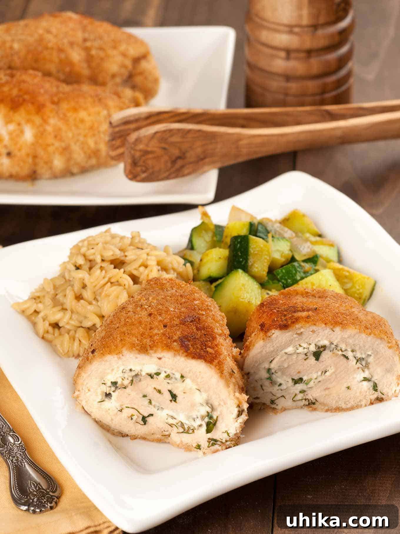 Gourmet Herbed Goat Cheese Stuffed Chicken 2 Rolled, crumb-coated chicken breasts filled with herbed goat cheese, served with sautéed zucchini and orzo on a square white plate, more chicken in the background.