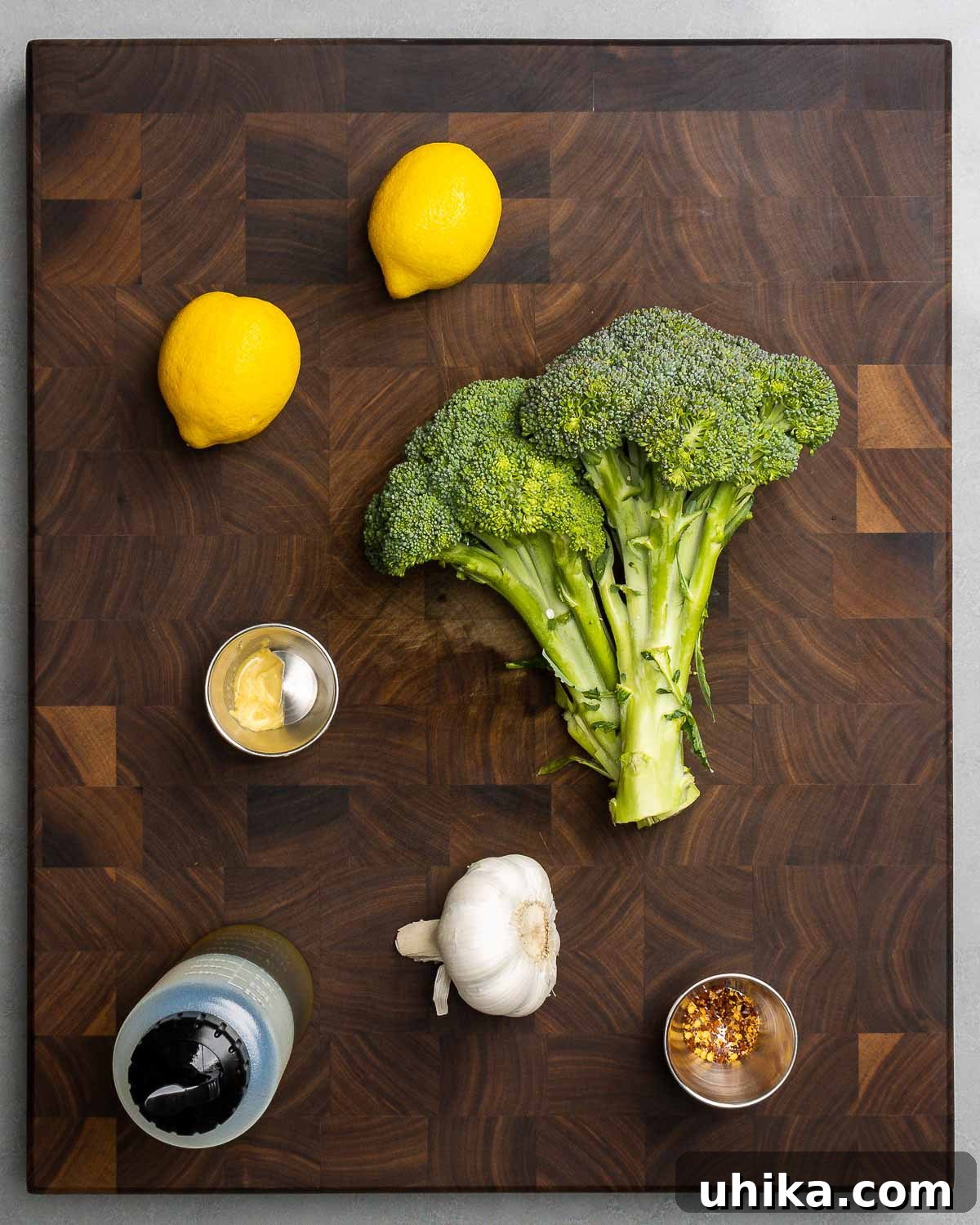 A visual display of all the fresh ingredients required for the Italian broccoli salad: bright green broccoli, radiant yellow lemons, a jar of Dijon mustard, clear extra virgin olive oil, fragrant garlic cloves, and a small pile of hot red pepper flakes.
