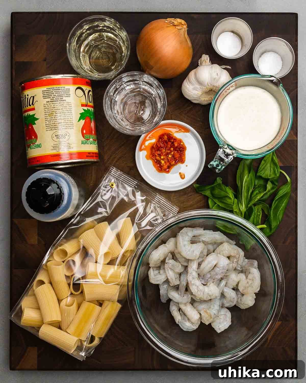 A flat lay photograph displaying all the fresh ingredients for Shrimp alla Vodka: vine-ripened tomatoes, a bottle of white wine, vodka, garlic cloves, salt, baking soda, Calabrian chili paste, heavy cream, olive oil, rigatoni pasta, and fresh basil, alongside a bowl of raw shrimp.