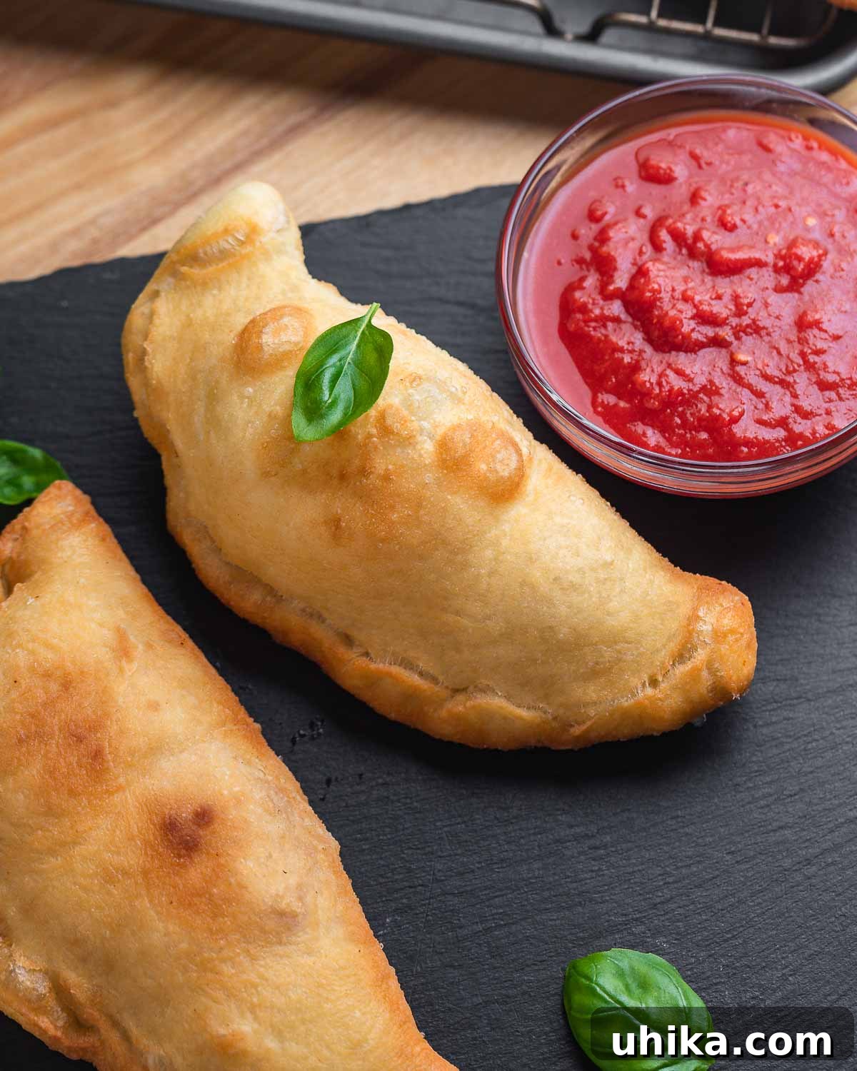 Crispy Italian Panzerotti 2 Two golden panzerotti resting on a black slate, garnished with fresh basil leaves and accompanied by a small bowl of vibrant marinara sauce.