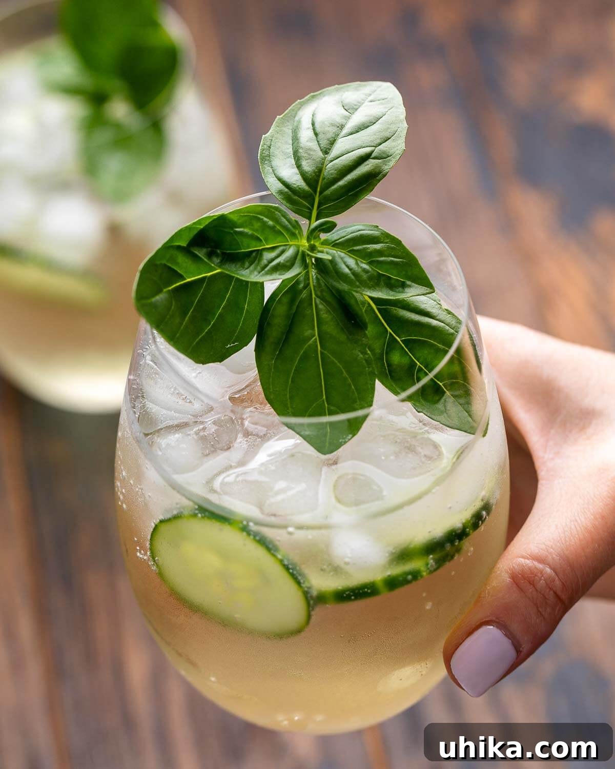 Effervescent Lillet Bliss 4 A hand gently holds a glass of Lillet Spritz, garnished with basil and cucumber, against a soft-focus wooden table background.