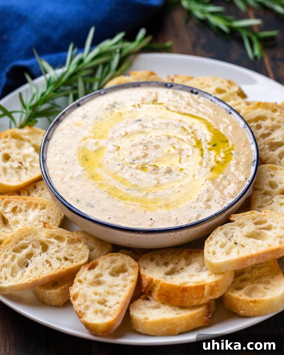 A bowl of creamy cannellini bean dip garnished with fresh rosemary and served with golden-brown crostini, perfect for an elegant appetizer.