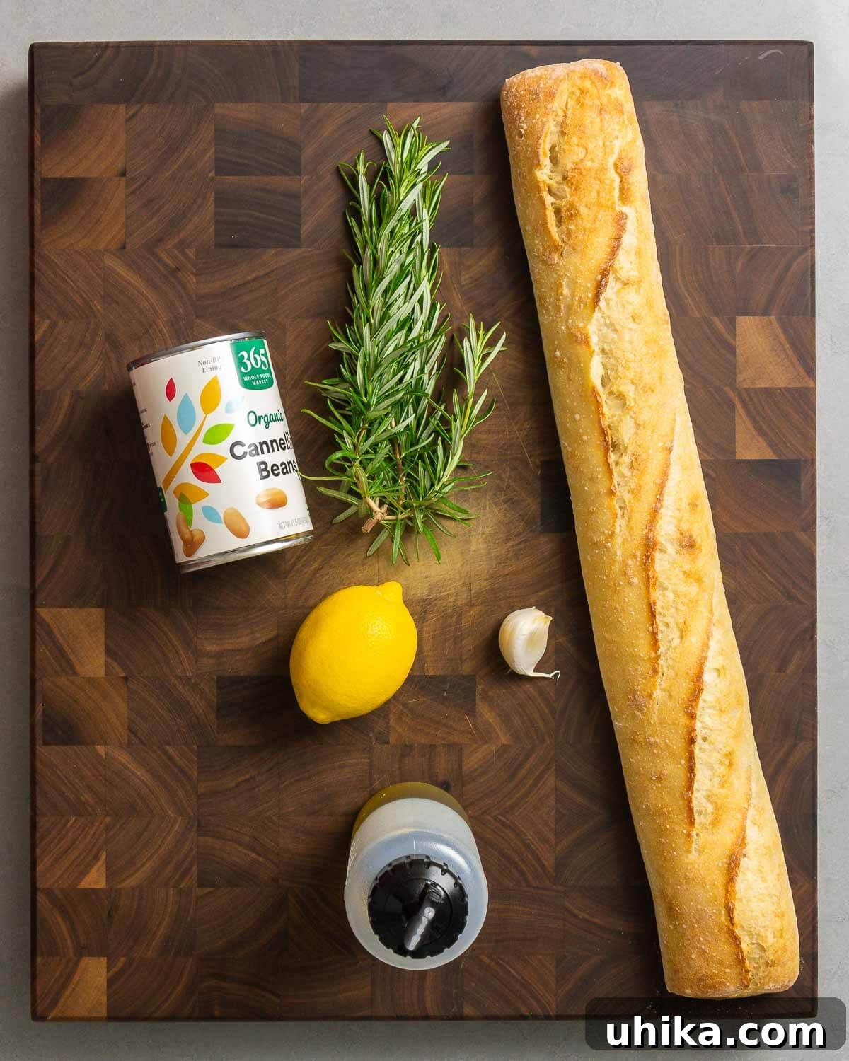Key ingredients for cannellini bean dip arranged on a rustic surface: cannellini beans, fresh rosemary sprigs, a whole lemon, garlic cloves, and a bottle of extra virgin olive oil.