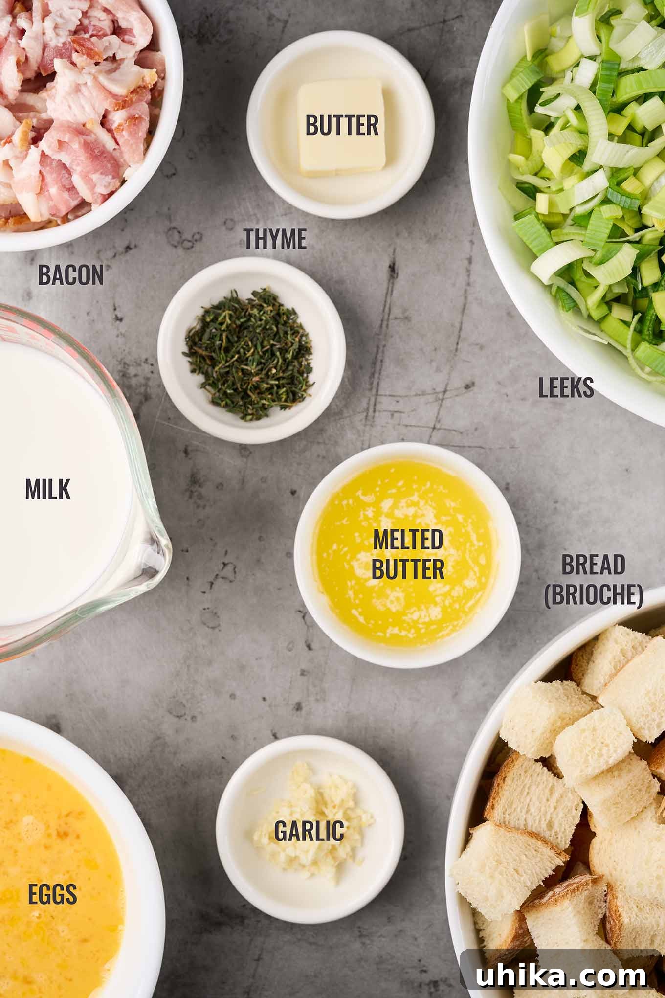 An overhead layout of key ingredients for savory bread pudding, featuring bread cubes, eggs, milk, fresh garlic, thick-cut bacon, sliced leeks, butter, and thyme sprigs.