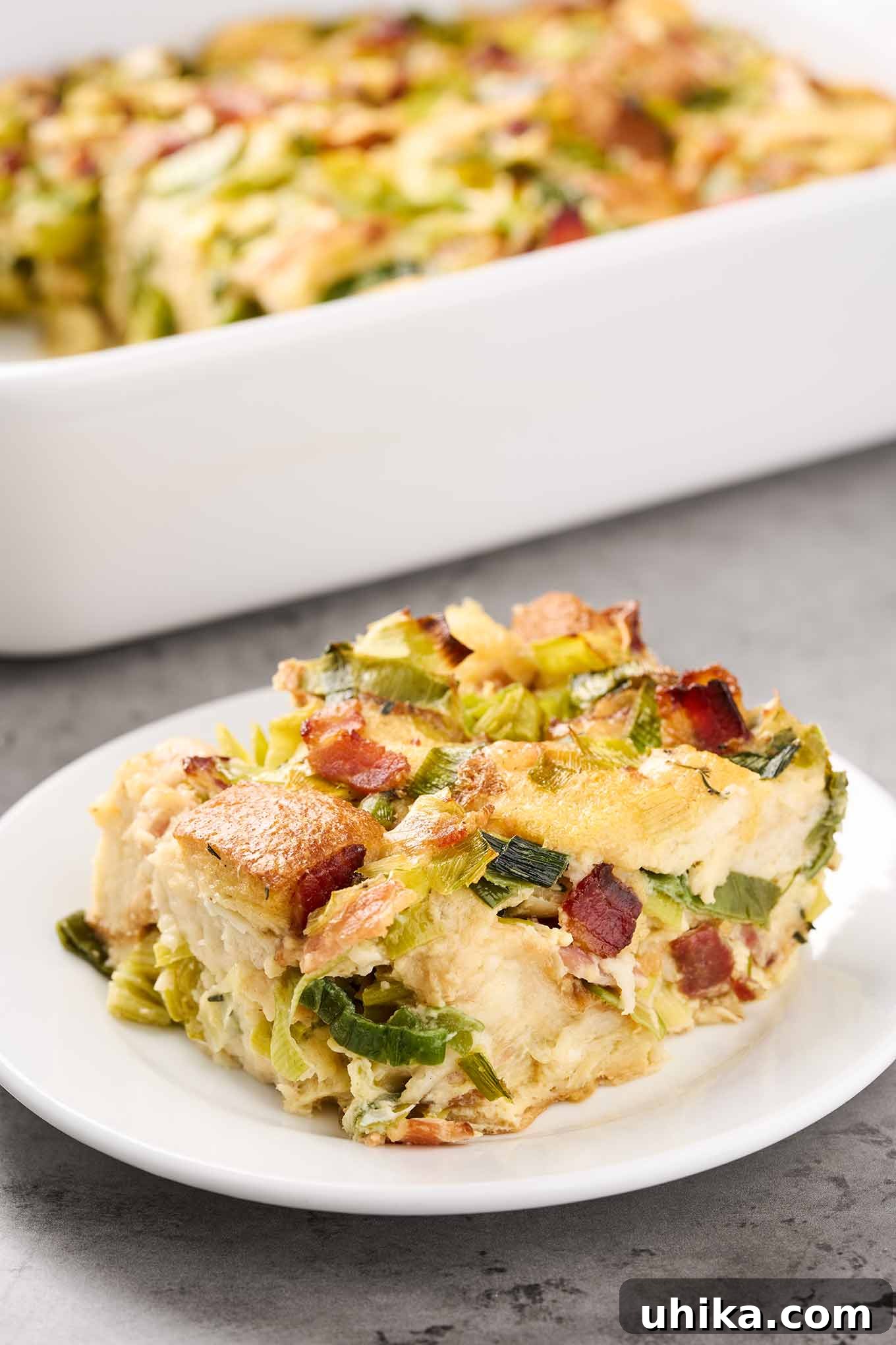 A close-up shot of a single serving of savory bread pudding on a white plate, showcasing its soft, custardy interior filled with crisp bacon pieces and tender sautéed leeks, with the baking dish blurred in the background.