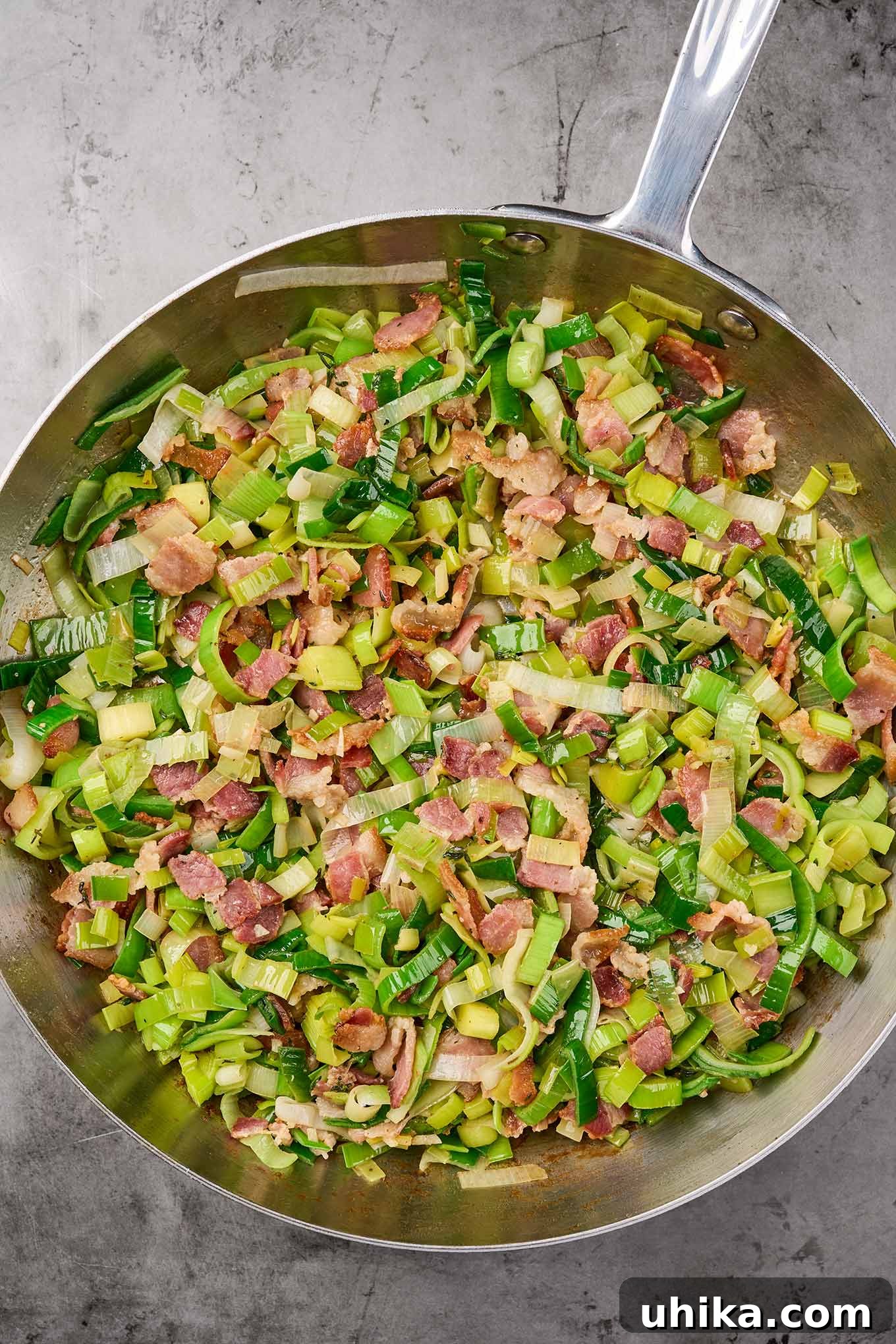 A stainless steel frying pan showcasing cooked thick-cut bacon and soft, sautéed leeks, seasoned with fresh thyme, ready to be incorporated into the bread pudding mixture.