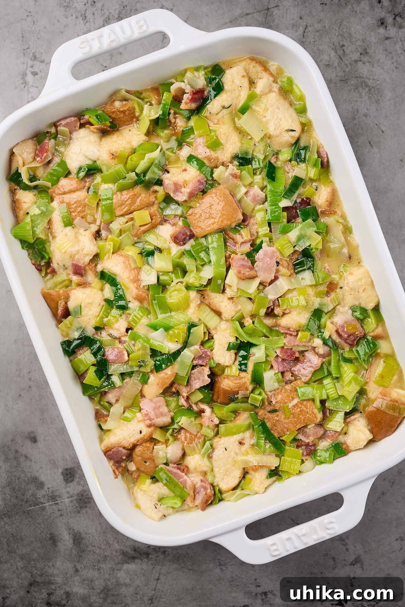 An assembled savory bread pudding with bacon and leeks in a baking dish, before it goes into the oven, showcasing the well-soaked bread and rich custard mixture.