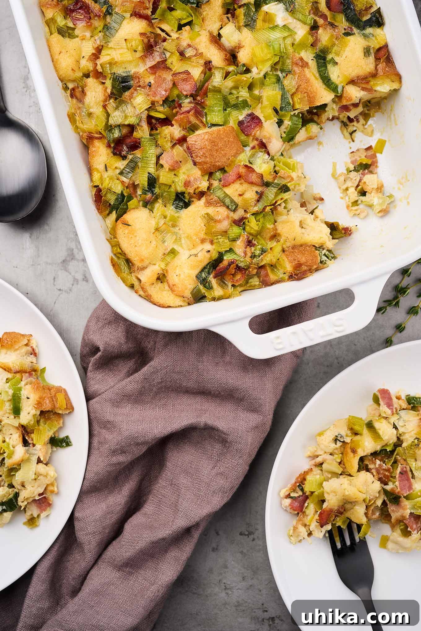A large casserole dish of savory bread pudding, with several servings already taken, positioned next to individual plates each holding a portion, garnished with fresh thyme sprigs, on a wooden surface.