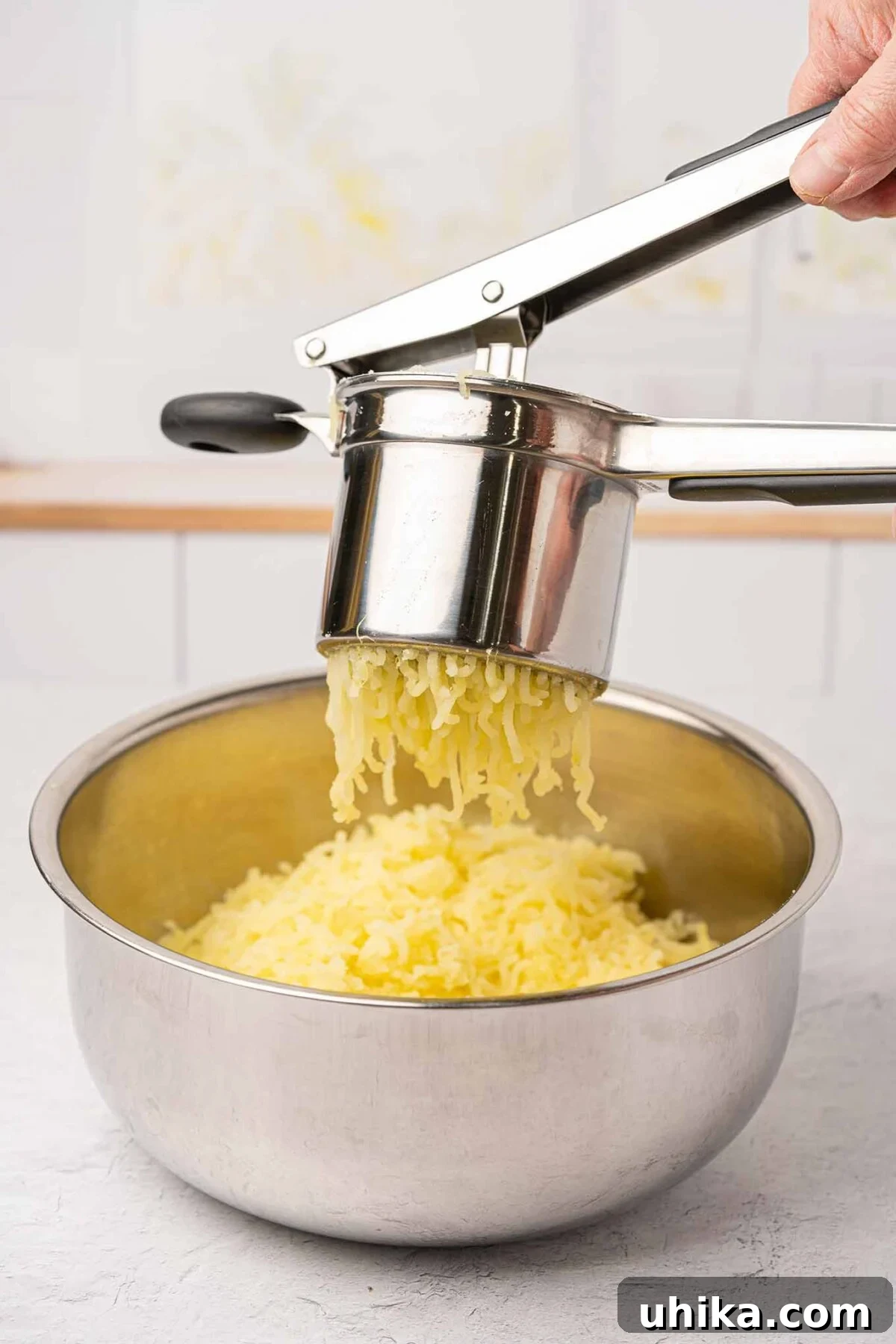 A detailed how-to photo capturing perfectly cooked potatoes being pressed through a potato ricer, demonstrating a key technique for achieving incredibly smooth and fluffy mashed potatoes.
