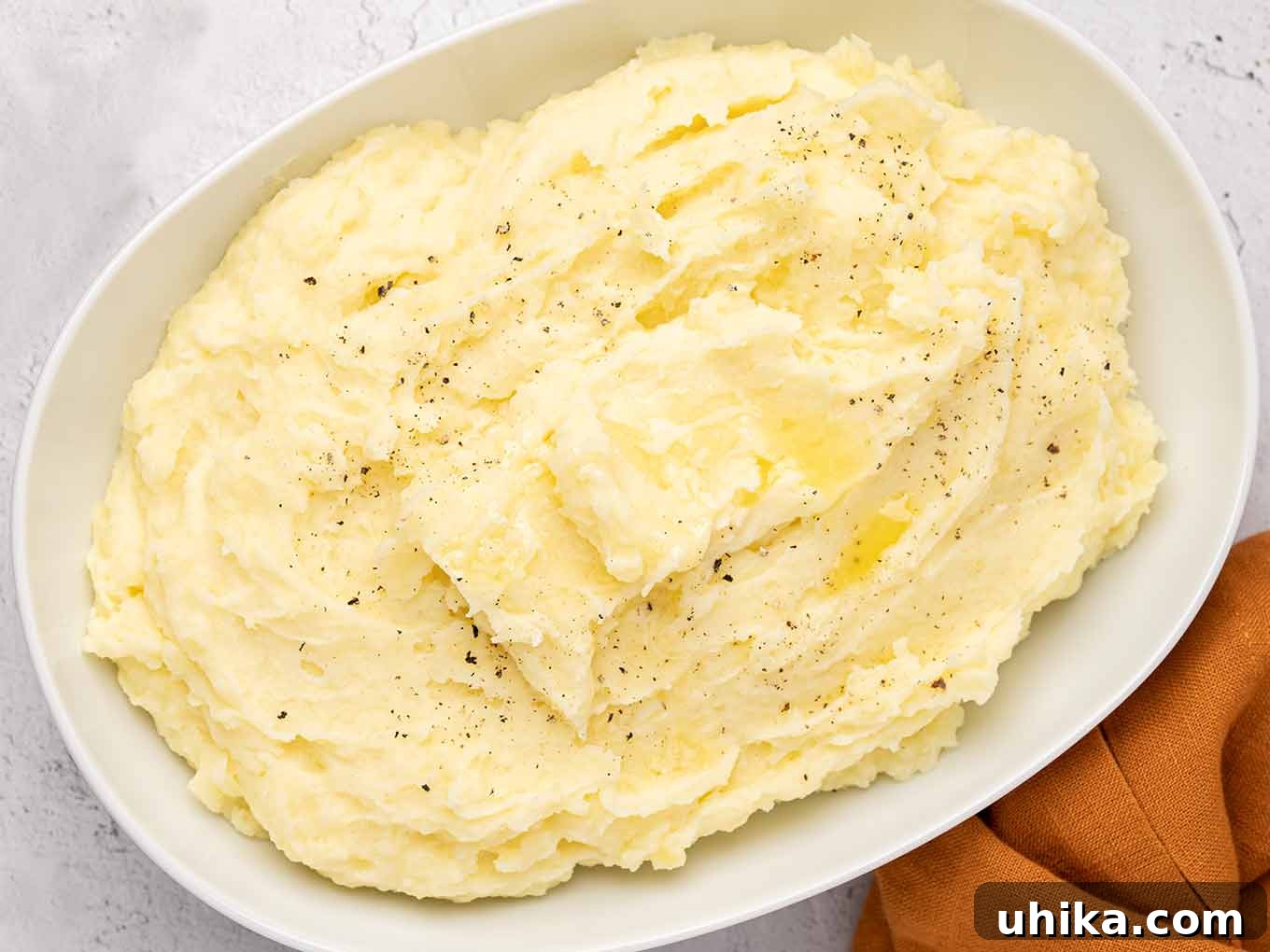 An inviting overhead view of a perfectly crafted serving bowl filled with creamy sour cream mashed potatoes, elegantly presented on a sophisticated textured gray background, with a vibrant rust-colored linen napkin adding a touch of warmth in the foreground.