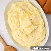 An oval serving bowl of creamy mashed potatoes with a melting pat of butter in the middle, a wooden spoon and linen napkin on either side of the bowl.