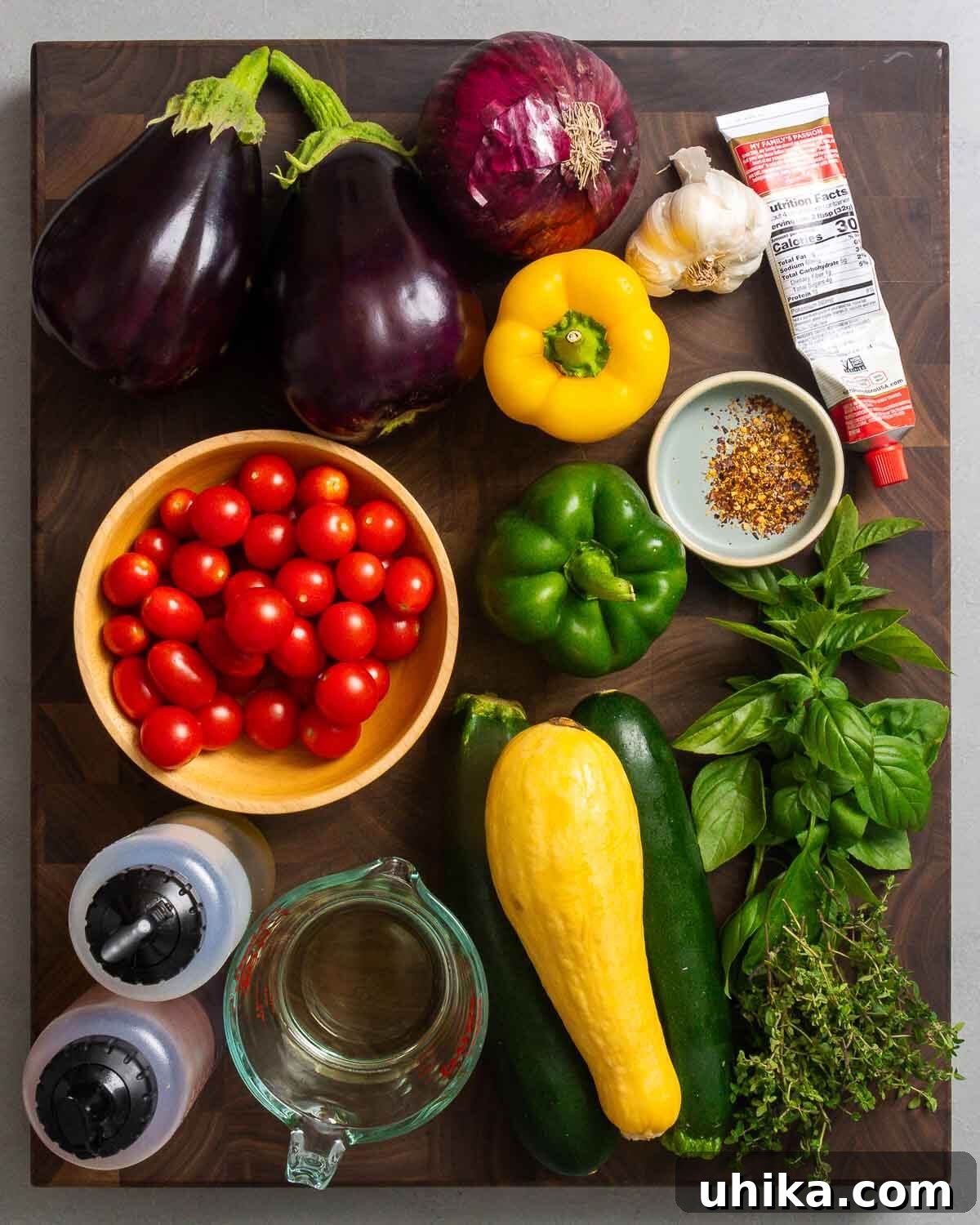 A selection of fresh Ratatouille ingredients laid out: eggplant, red onion, garlic cloves, tomato paste, vibrant bell peppers, hot red pepper flakes, ripe tomatoes, yellow squash, green zucchini, olive oil, vinegar, white wine, fresh basil, and thyme sprigs.