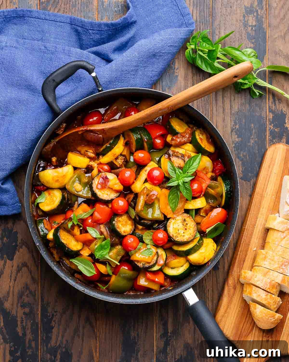 Overhead shot of a beautifully cooked Ratatouille in a black pan, garnished with fresh basil leaves, with a side of crusty baguette slices.