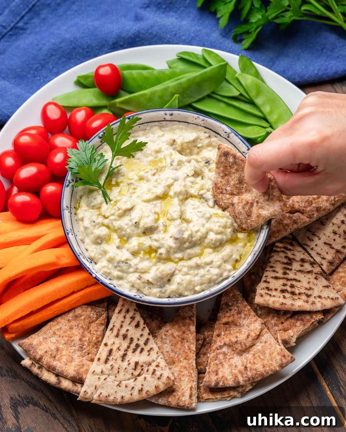 Overhead shot of baba ganoush platter with hands dipping pita into bowl, garnished with olive oil and fresh herbs.