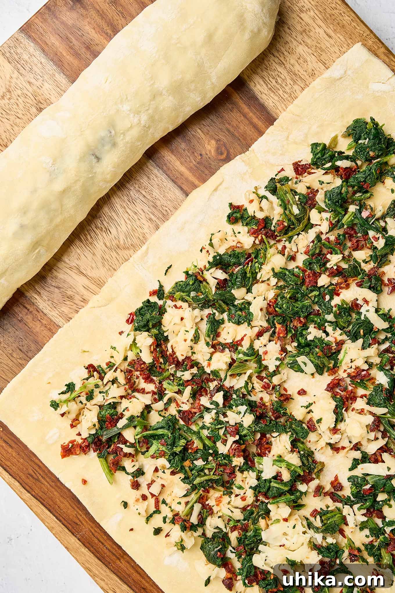 Savory Spinach Swirls 5 Puff pastry sheet topped with an even layer of spinach, sun-dried tomatoes, and cheese filling, shown next to an already rolled pastry log, demonstrating the preparation process.