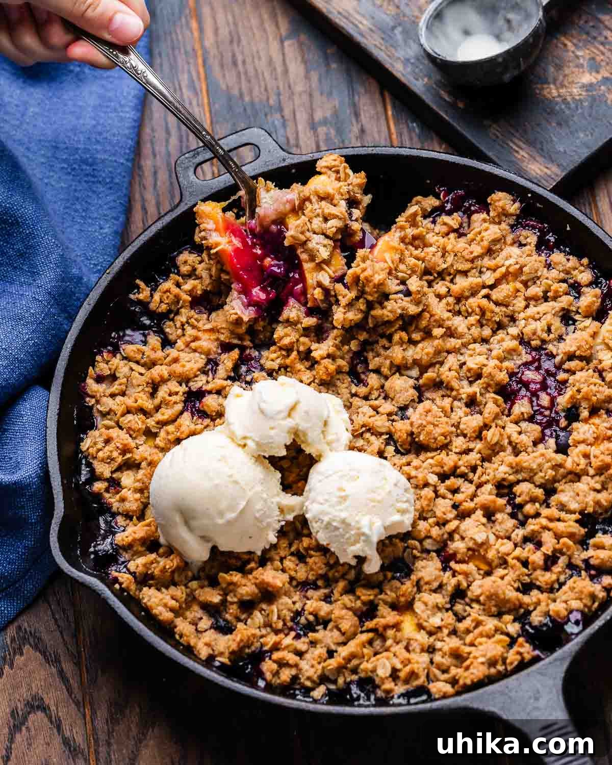 Sun-Kissed Peach and Blueberry Crumble 7 Peach blueberry crisp in cast iron pan on table with spoon scooping out some of it, emphasizing its deliciousness.