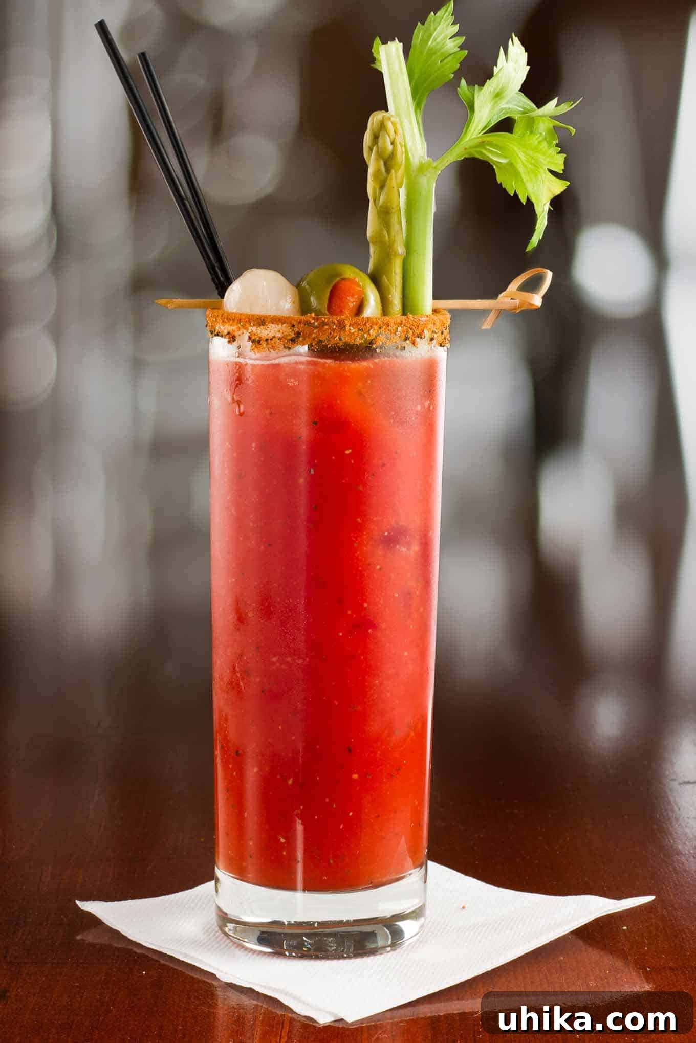 Zesty Red Reviver 2 Tall glass of Bloody Mary cocktail garnished with celery stalk, pickled asparagus, green olive, and cocktail onion, served with a spiced rim and black straw.