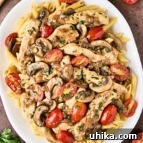 An overhead view of chicken Marsala pasta on a white oval serving platter, surrounded by a scattering of halved grape tomatoes, a sprig of parsley, and a wooden spoon.