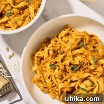Two bowls of creamy pumpkin pasta with pancetta and sage served in white bowls, a glass of white wine and cheese grater in the background.