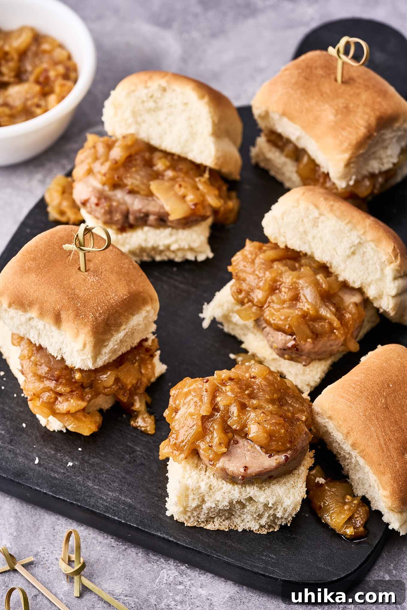 Maple Mustard Glazed Pork Tenderloin Sliders 2 Pork tenderloin slider sandwiches secured with bamboo skewers on a serving board with a bowl of caramelized onion topping in the background, showcasing a perfect appetizer for any gathering.