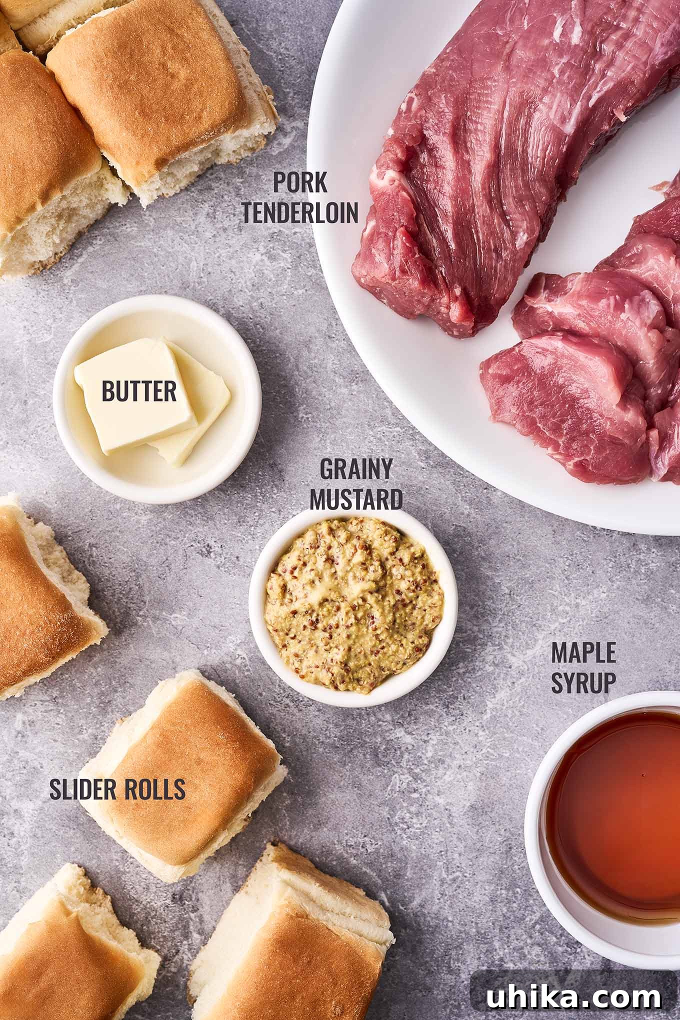 Maple Mustard Glazed Pork Tenderloin Sliders 3 An overhead shot showing all essential ingredients for making delicious pork tenderloin sliders, including a whole pork tenderloin, soft slider buns, pure maple syrup, robust grainy mustard, and creamy butter, neatly arranged on a white surface.