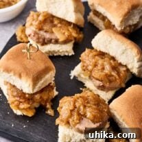 Pork tenderloin slider sandwiches secured with bamboo skewers on a serving board with a bowl of caramelized onion topping in the background.