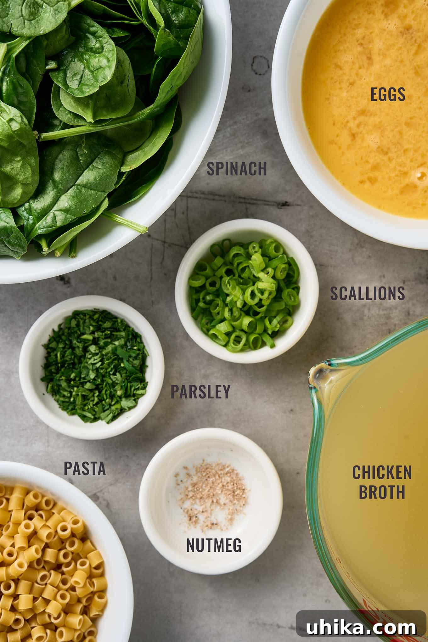 Silken Stracciatella with Fresh Spinach and Tender Pasta 3 Labeled overhead view of ingredients for stracciatella soup, including spinach, eggs, scallions, parsley, pasta, nutmeg, and chicken broth.