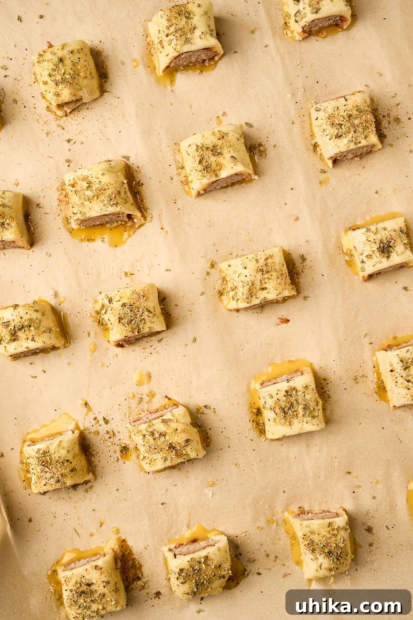 Golden Sausage Puffs 7 Prepared puff pastry sausage rolls, already sliced into individual pieces, glistening with an egg wash, and delicately sprinkled with dried oregano and all-purpose seasoning, arranged on a baking sheet.