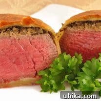 A dinner plate with an individual beef Wellington, sliced in half to show the perfectly cooked beef, topping of mushroom duxelles, and flaky pastry wrap.