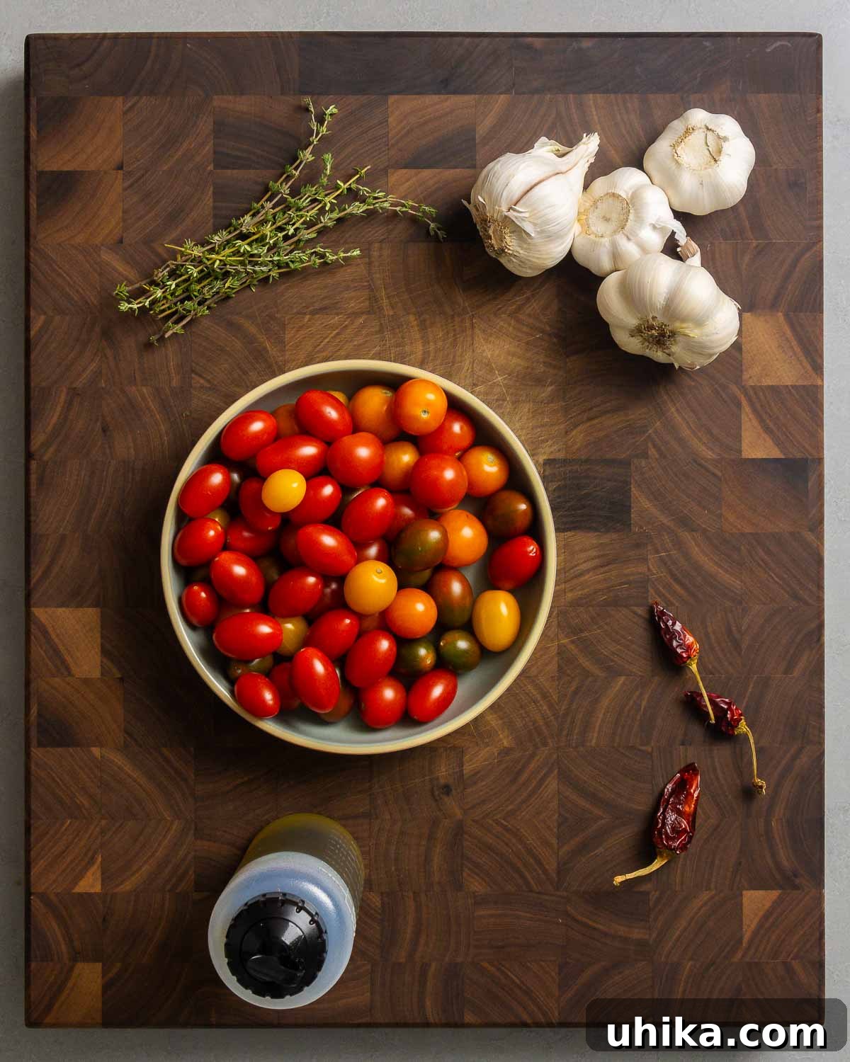 Rich Roasted Tomato Jewels 3 Fresh ingredients laid out for tomato garlic confit: thyme, garlic cloves, mixed cherry and grape tomatoes, dried red chili peppers, and a bottle of olive oil.