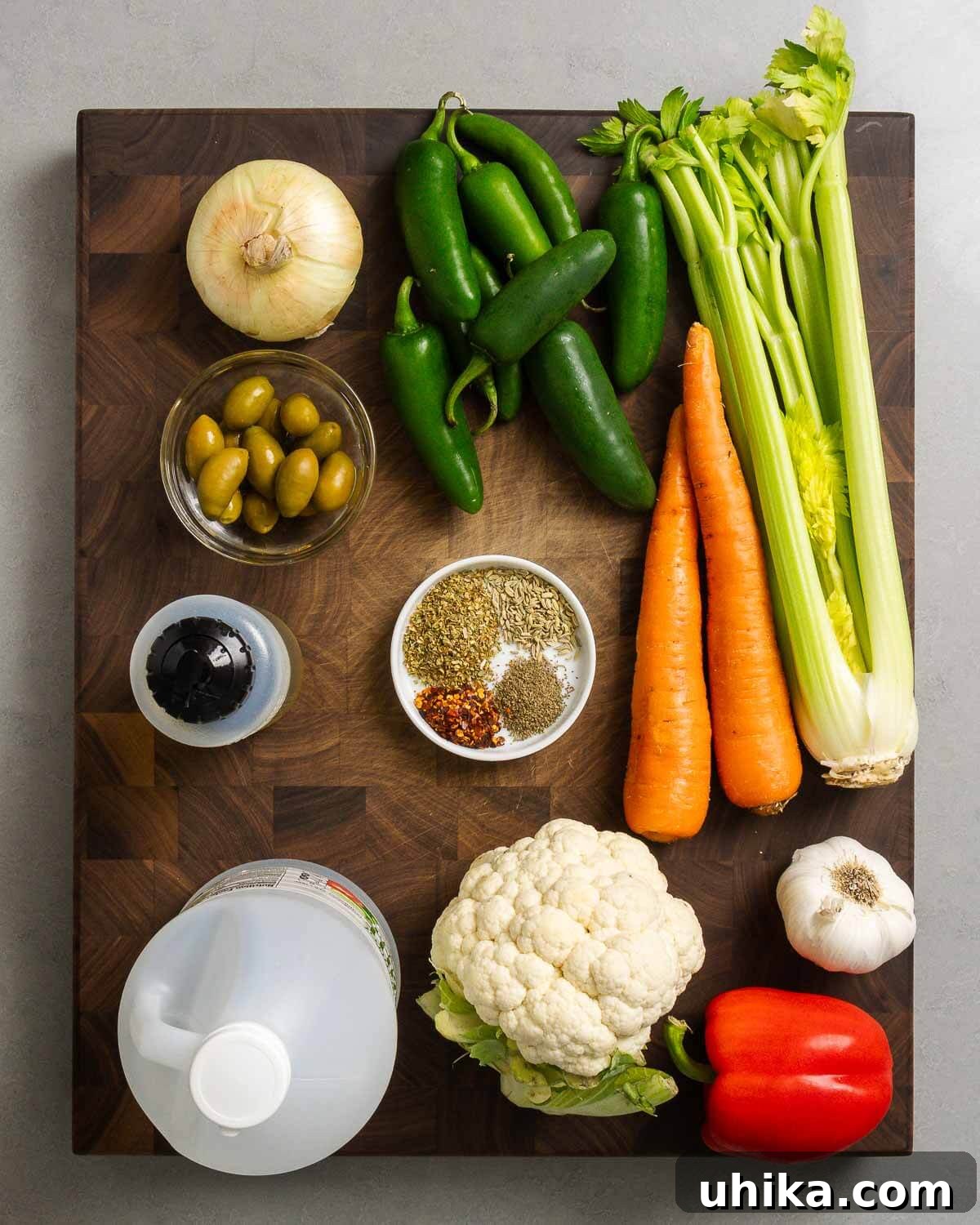 Zesty Garden Relish 3 A flat lay image displaying all the raw ingredients required for making giardiniera, including a sweet onion, hot peppers, carrots, celery, green olives, olive oil, various spices, white vinegar, cauliflower, garlic, and a red bell pepper.