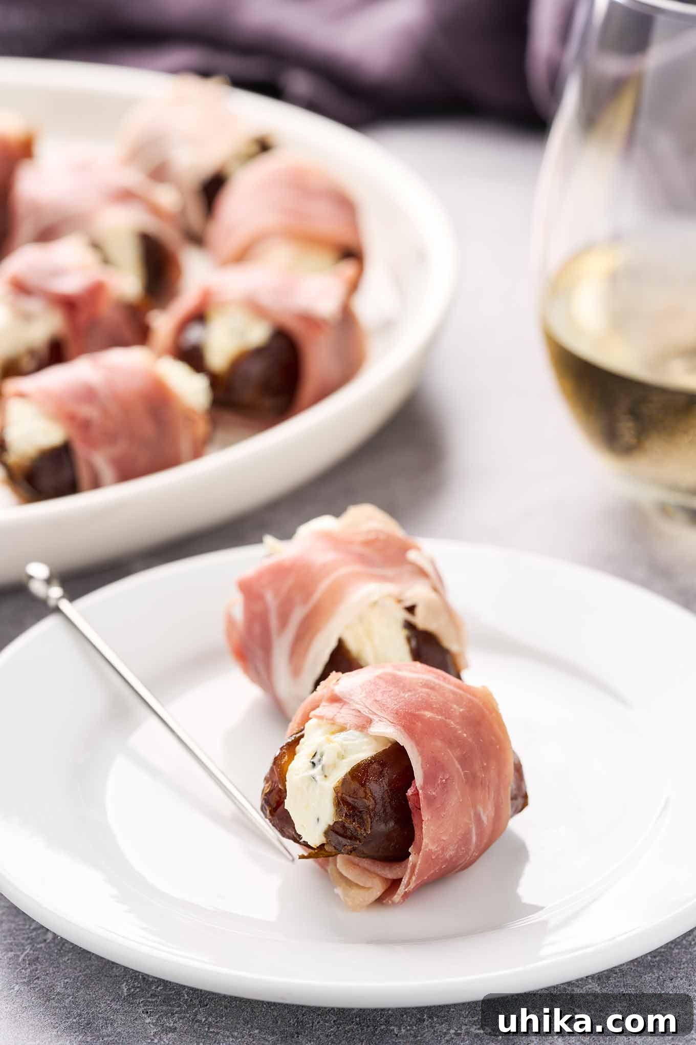 Sweet Salty Creamy Prosciutto Date Bites 2 Stuffed dates wrapped in prosciutto on an appetizer plate with a silver pick, a glass of white wine and serving plate in the background.