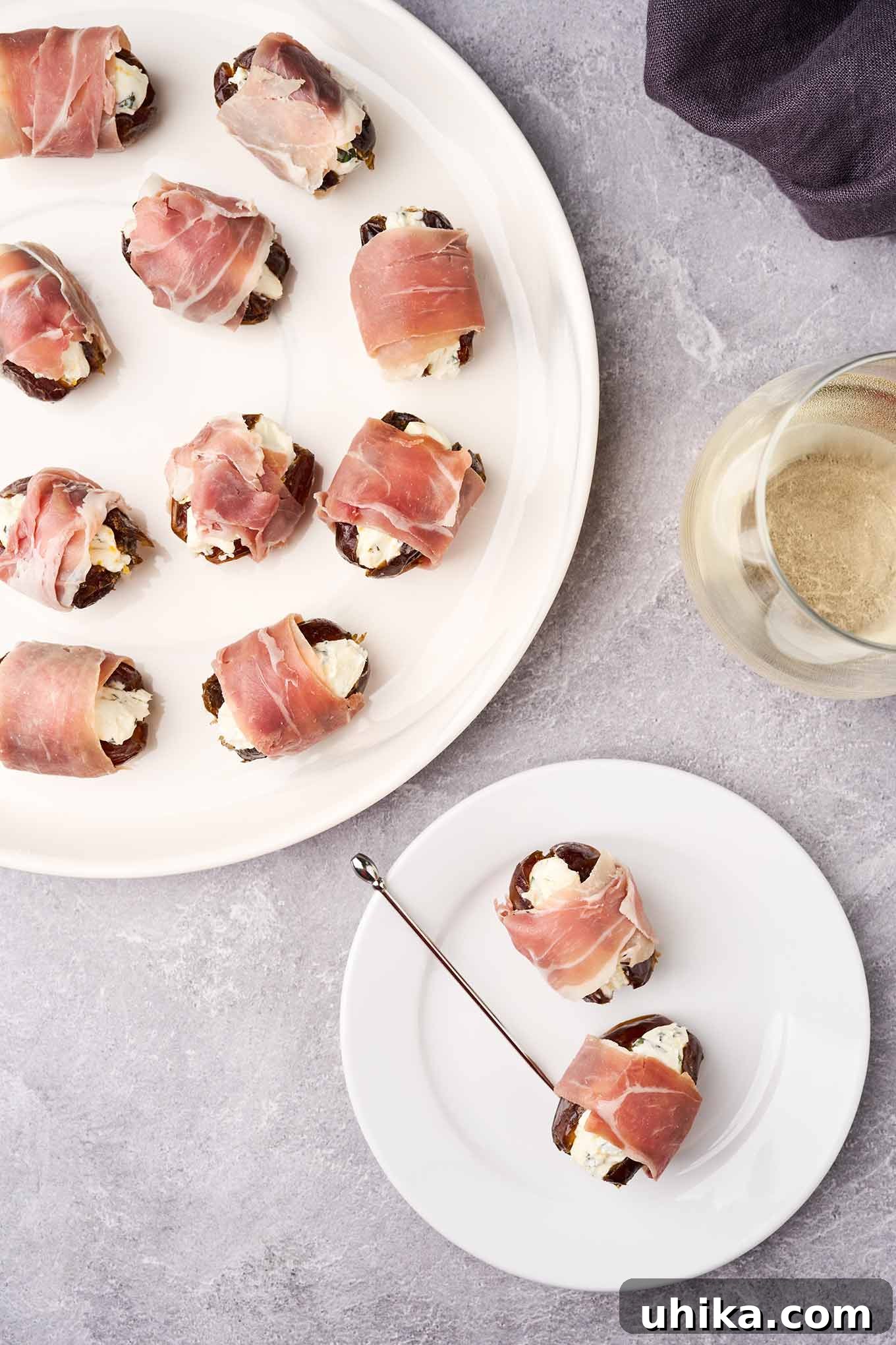 Sweet Salty Creamy Prosciutto Date Bites 5 An overhead view of a serving plate of prosciutto-wrapped stuffed dates, a glass of white wine, and an appetizer plate with two stuffed dates and a silver cocktail pick.