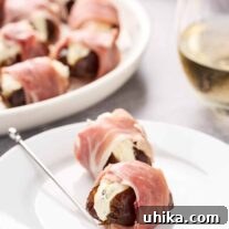 Stuffed dates wrapped in prosciutto on an appetizer plate with a silver pick, a glass of white wine and serving plate in the background.