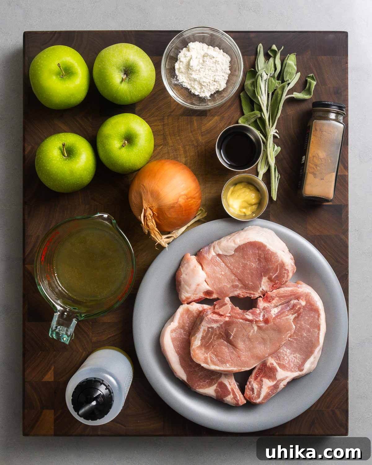 All ingredients for the pork chops with apples recipe laid out: apples, flour, fresh sage, balsamic vinegar, ground cinnamon, Dijon mustard, sliced onion, chicken stock, raw pork chops, and olive oil.