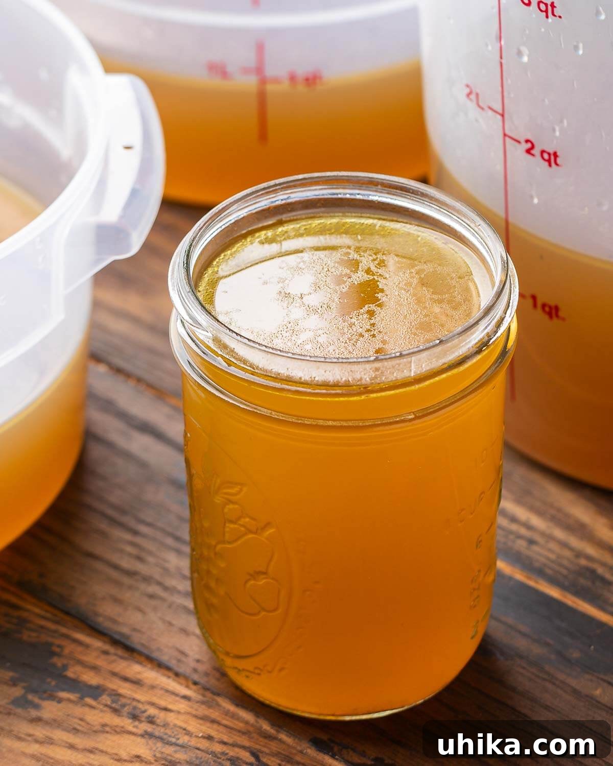 Liquid Gold Beef Stock 2 Mason jar filled with rich, golden brown beef stock along with three large storage containers in the background, showcasing abundant homemade stock.