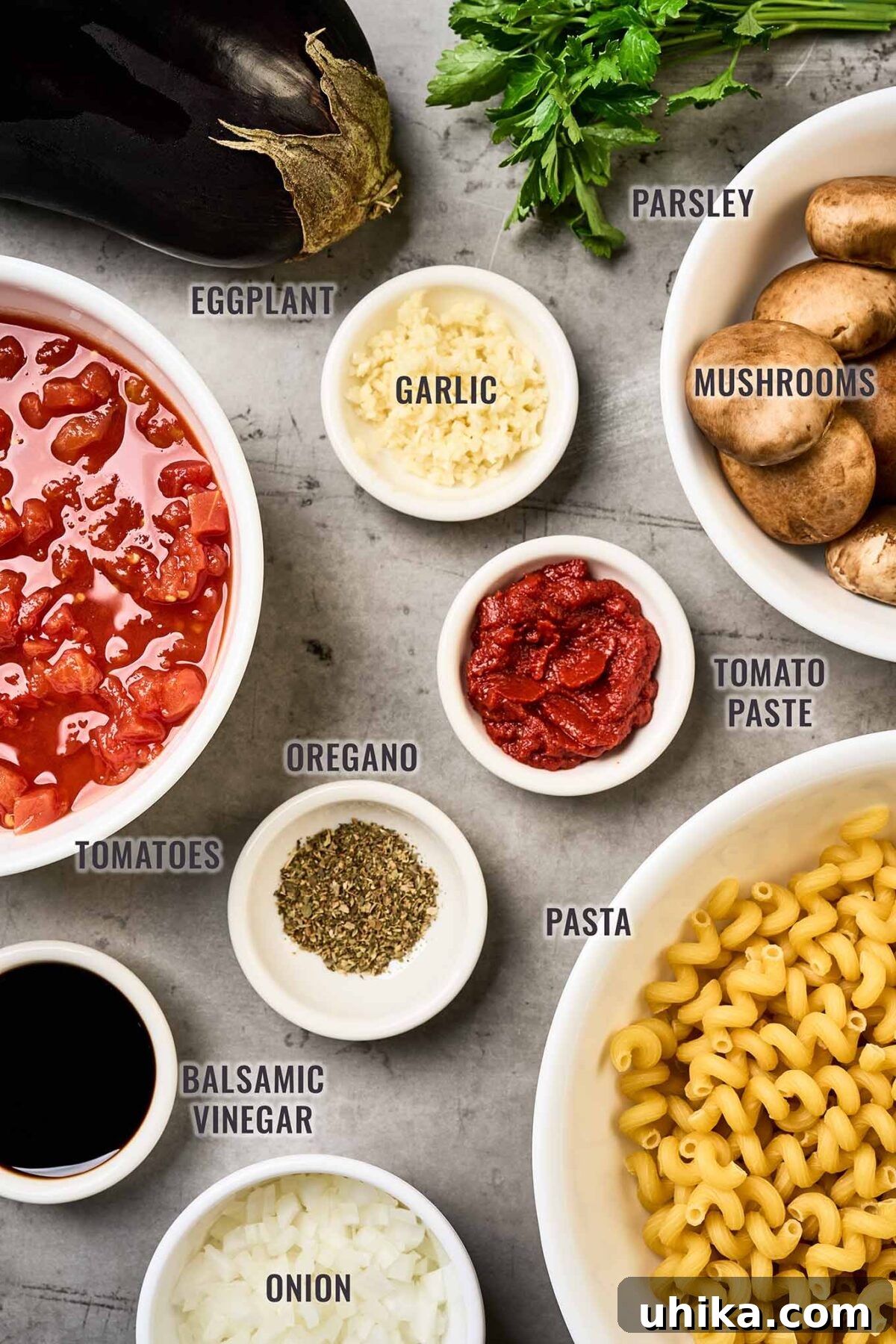 Savory Eggplant Mushroom Pasta 3 A labeled, overhead view of the ingredients needed to make eggplant and mushroom pasta, including eggplant, pasta, mushrooms, onion, garlic, parsley, tomato paste, oregano, balsamic vinegar, and canned tomatoes.