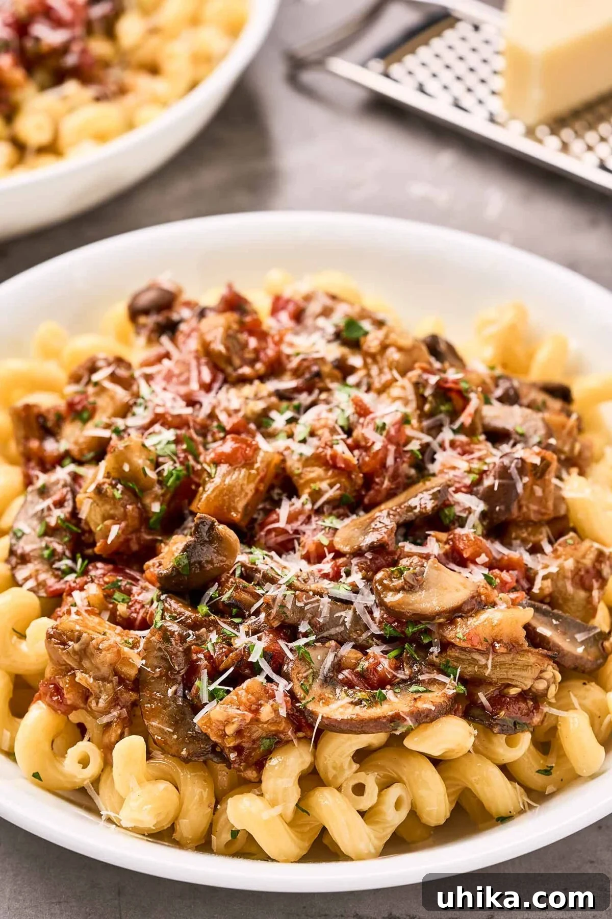 Savory Eggplant Mushroom Pasta 7 A single serving bowl of eggplant and mushroom pasta topped with grated Parmesan cheese and parsley, with a second blurred bowl and a cheese grater in the background.